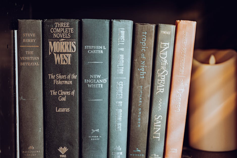 A row of novels by various authors standing upright on a bookshelf next to an unlit pillar candle.