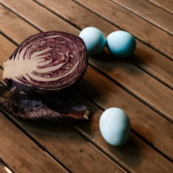 A halved red cabbage, a loose cabbage leaf, and three blue eggs are arranged on a wooden slatted surface.