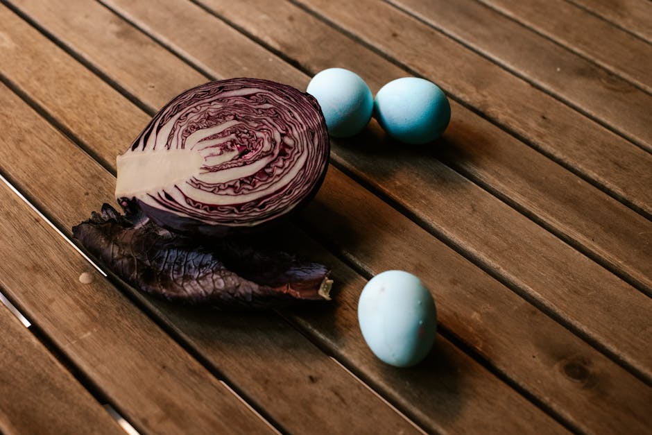 A halved red cabbage, a loose cabbage leaf, and three blue eggs are arranged on a wooden slatted surface.