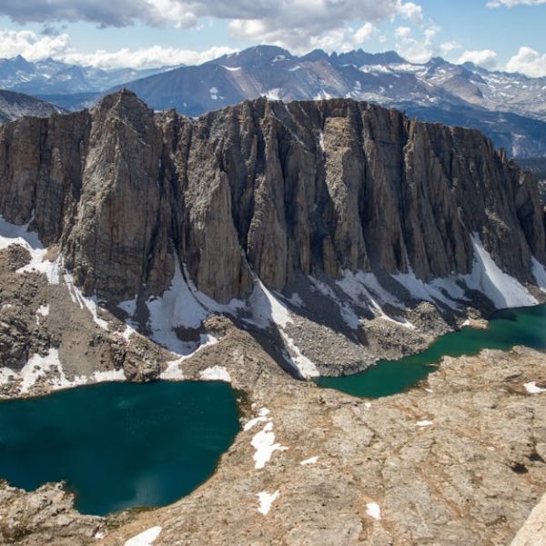 Rocky cliffs rise above alpine lakes with patches of snow, set against a backdrop of distant mountains under a partly cloudy sky.