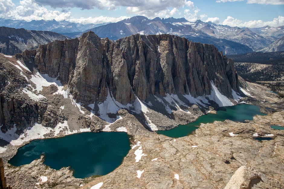 Rocky cliffs rise above alpine lakes with patches of snow, set against a backdrop of distant mountains under a partly cloudy sky.