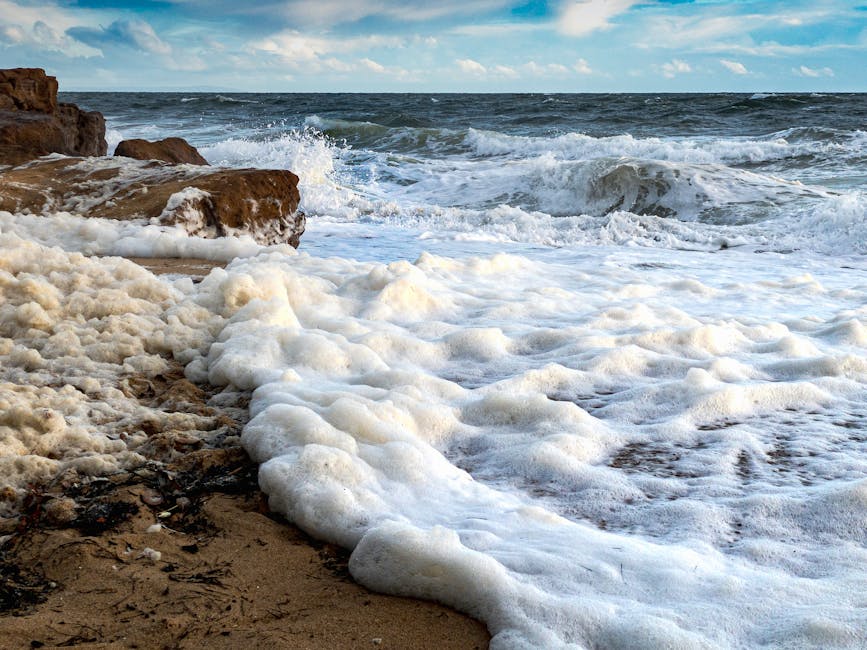 Waves crash onto a sandy beach, creating sea foam along the shoreline, with rocky outcrops and a partly cloudy sky in the background.