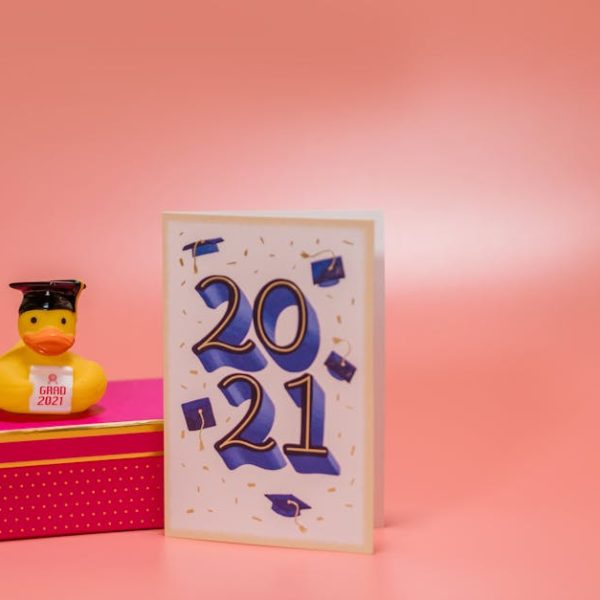 A rubber duck with a graduation cap and "Grad 2021" sign sits on a pink box next to a card that reads "2021" with graduation cap graphics, against a pink background.