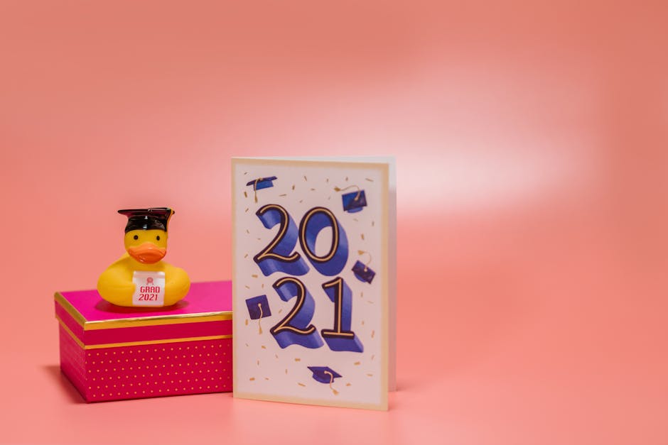 A rubber duck with a graduation cap and "Grad 2021" sign sits on a pink box next to a card that reads "2021" with graduation cap graphics, against a pink background.