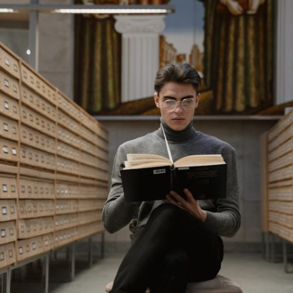 A person wearing glasses and a gray turtleneck sits between rows of card catalog drawers, reading a large book in a library.
