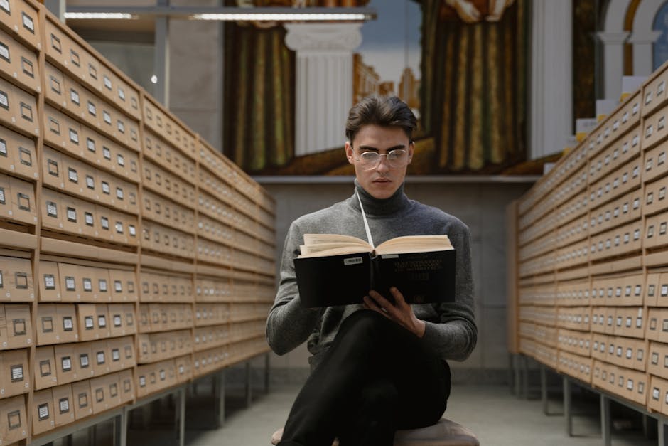 A person wearing glasses and a gray turtleneck sits between rows of card catalog drawers, reading a large book in a library.