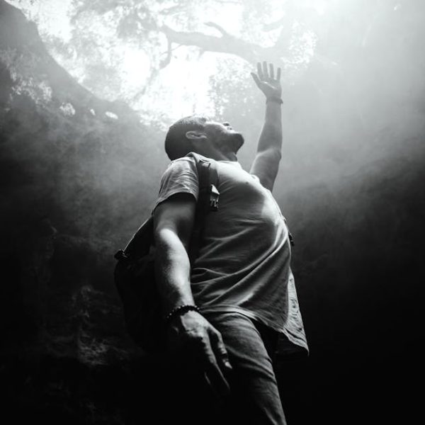 A man with a backpack stands at the bottom of a cave or pit, reaching upward toward the light above.