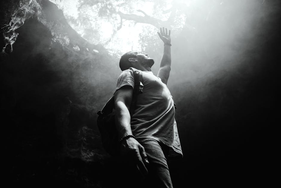 A man with a backpack stands at the bottom of a cave or pit, reaching upward toward the light above.