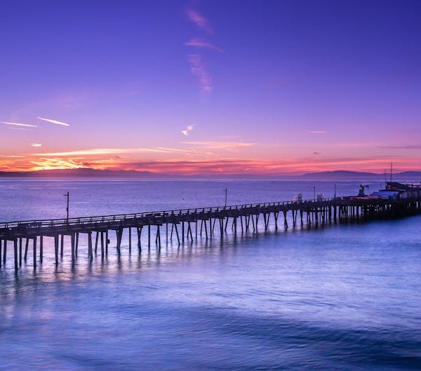 A long wooden pier extends over calm water at sunset, with a colorful sky featuring shades of purple, orange, and blue.