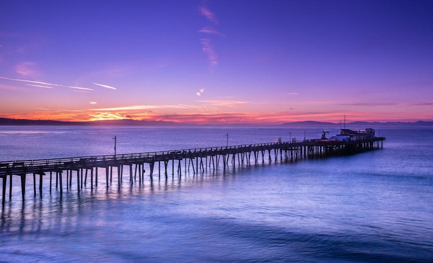 A long wooden pier extends over calm water at sunset, with a colorful sky featuring shades of purple, orange, and blue.