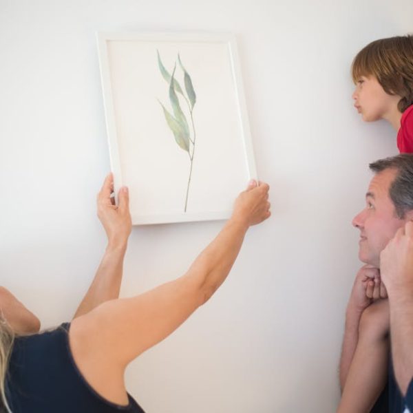 A woman hangs a framed botanical print on a white wall while a man holds a child on his shoulders, all looking at the picture.