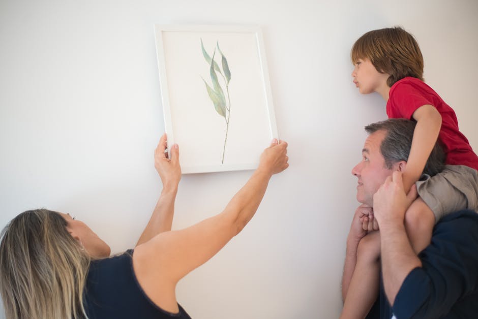 A woman hangs a framed botanical print on a white wall while a man holds a child on his shoulders, all looking at the picture.