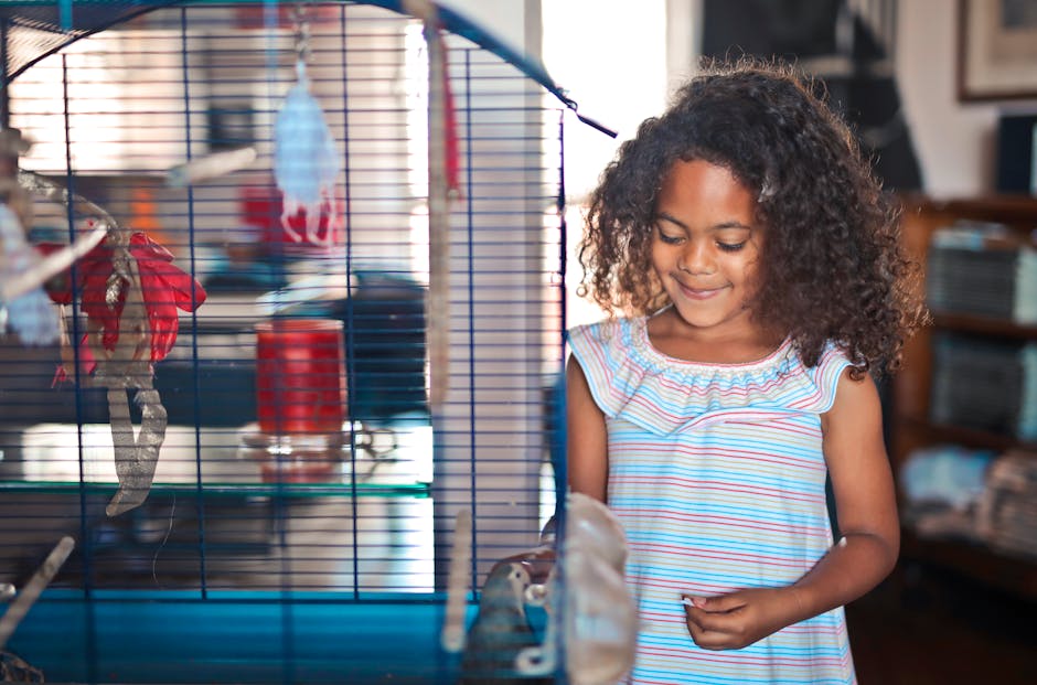 A young girl with curly hair stands next to a birdcage, smiling and looking at a small animal inside.