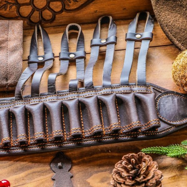 A brown leather ammunition belt with multiple loops lies on a wooden surface, surrounded by a hat, pinecone, evergreen branch, and decorative items.