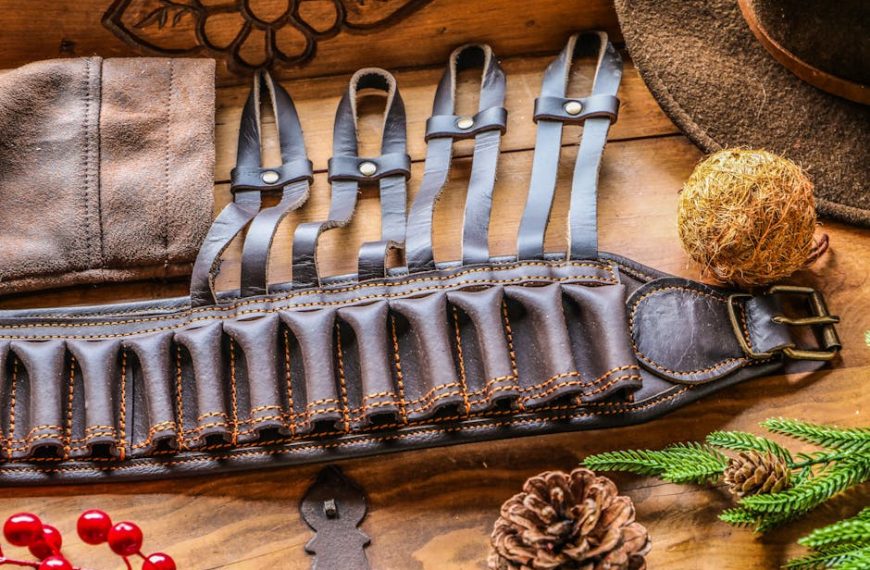 A brown leather ammunition belt with multiple loops lies on a wooden surface, surrounded by a hat, pinecone, evergreen branch, and decorative items.