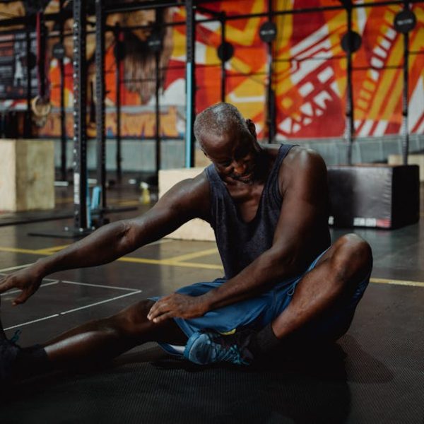 A man sits on a gym floor stretching his leg, touching his foot with one hand. Exercise equipment and a colorful mural are visible in the background. A water bottle is nearby.