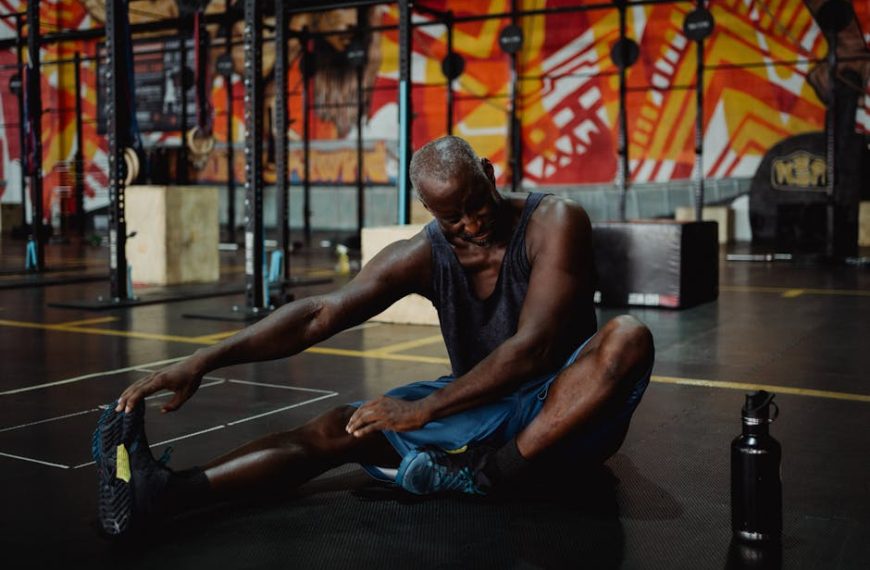 A man sits on a gym floor stretching his leg, touching his foot with one hand. Exercise equipment and a colorful mural are visible in the background. A water bottle is nearby.