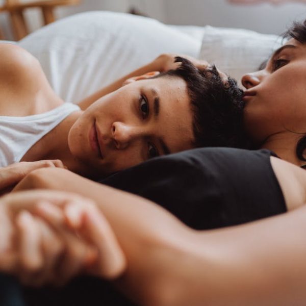 Two people are lying close together on a bed, one looking forward while the other rests their face near the first person's head.