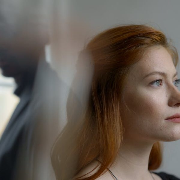 A woman with red hair looks ahead thoughtfully while a man stands blurred in the background near a window.