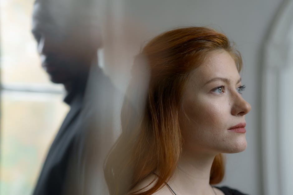A woman with red hair looks ahead thoughtfully while a man stands blurred in the background near a window.