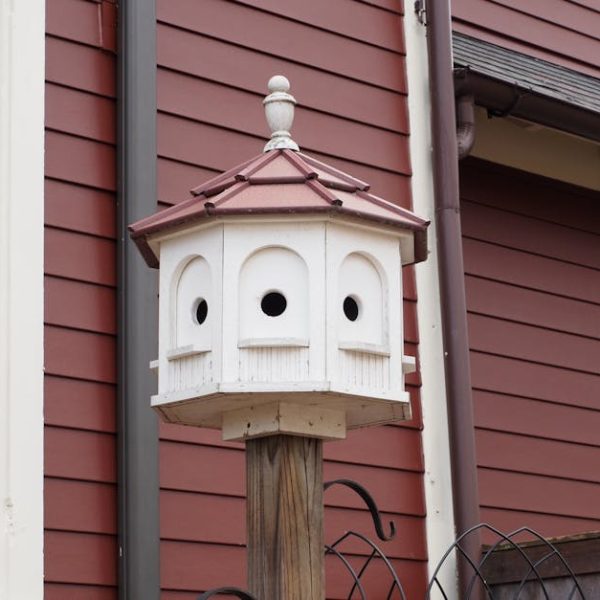 A white multi-compartment birdhouse with a red roof is mounted on a wooden post in front of a red house with cream trim.