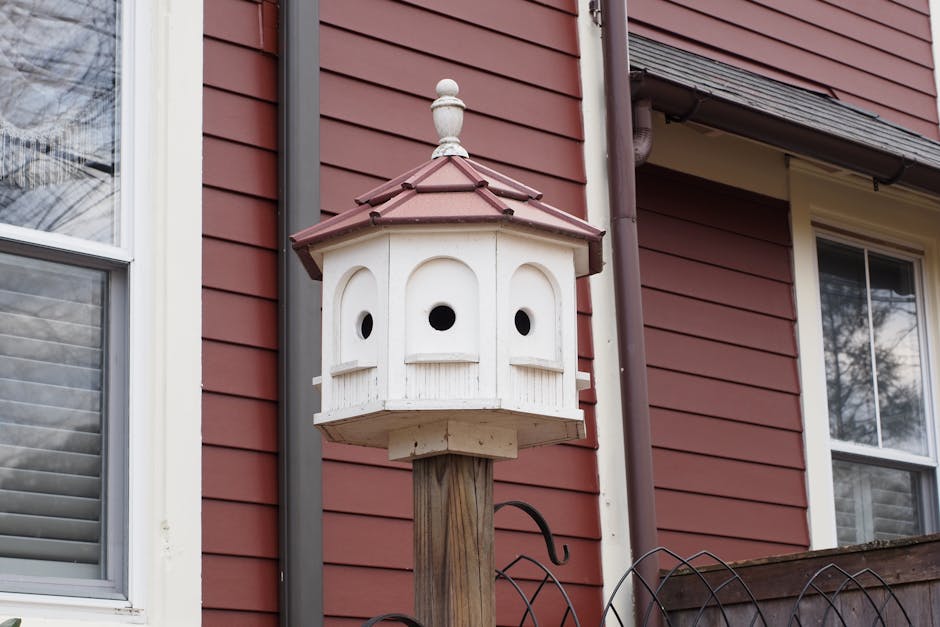 A white multi-compartment birdhouse with a red roof is mounted on a wooden post in front of a red house with cream trim.