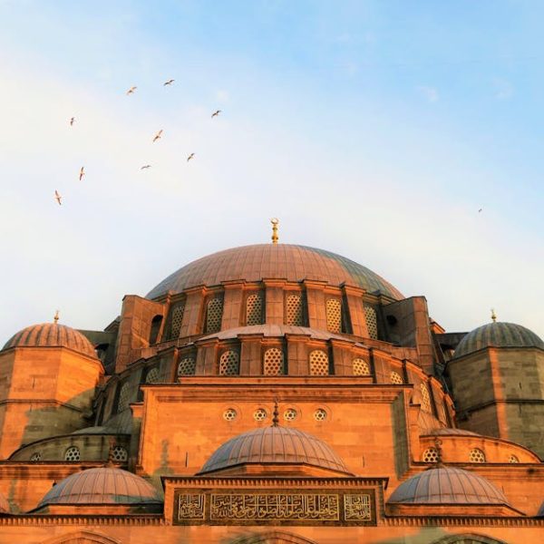 A large stone mosque with multiple domes, intricate windows, and Arabic script, with birds flying in the sky above at sunset.