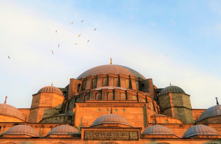 A large stone mosque with multiple domes, intricate windows, and Arabic script, with birds flying in the sky above at sunset.