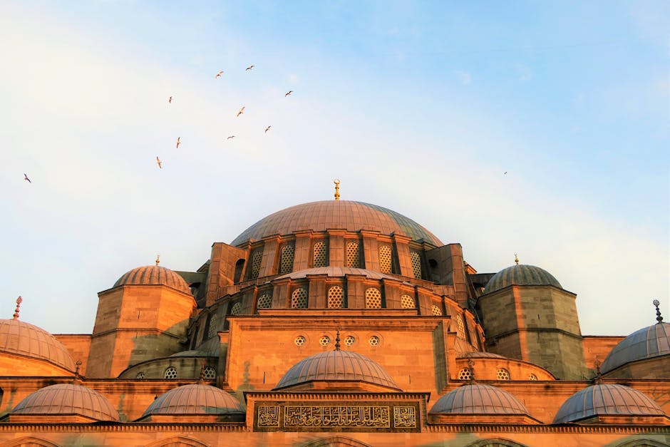 A large stone mosque with multiple domes, intricate windows, and Arabic script, with birds flying in the sky above at sunset.