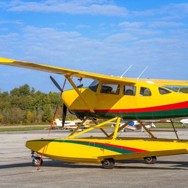 A yellow floatplane with green and red stripes is parked on an airport tarmac with trees and small planes in the background.