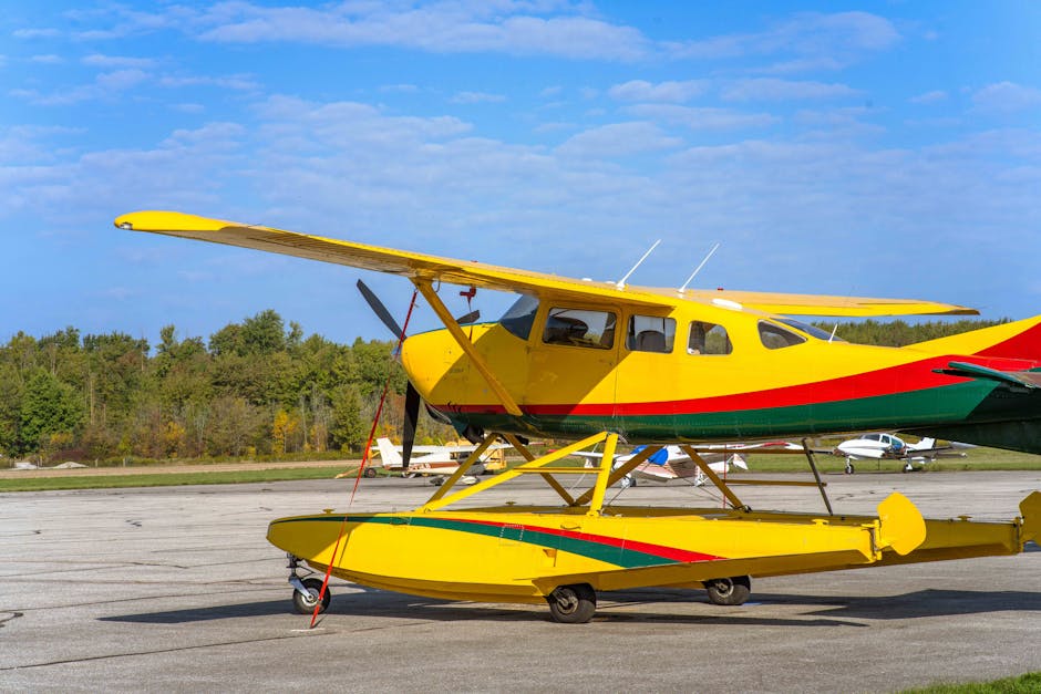 A yellow floatplane with green and red stripes is parked on an airport tarmac with trees and small planes in the background.