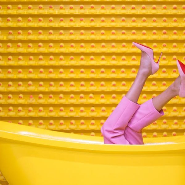 A person wearing pink pants and pink high heels lies in a yellow bathtub with legs sticking up against a yellow wall with a repeating pattern.