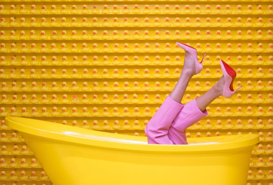 A person wearing pink pants and pink high heels lies in a yellow bathtub with legs sticking up against a yellow wall with a repeating pattern.